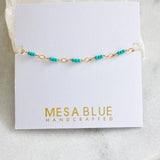 The Turquoise Seed Bead Bracelet by Mesa Blue features a delicate 18K gold plated chain adorned with small turquoise beads and includes an extender chain for the perfect fit, displayed on a white card labeled “Mesa Blue Handcrafted.”.
