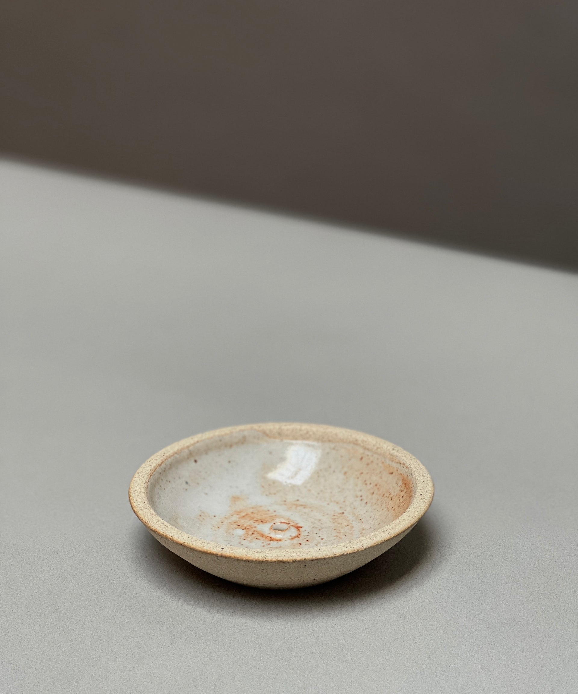 The Stoneware Holder | Incausa by Incausa is a small, shallow hand-thrown incense burner with a light beige and speckled glaze, displayed on a smooth, light gray surface.