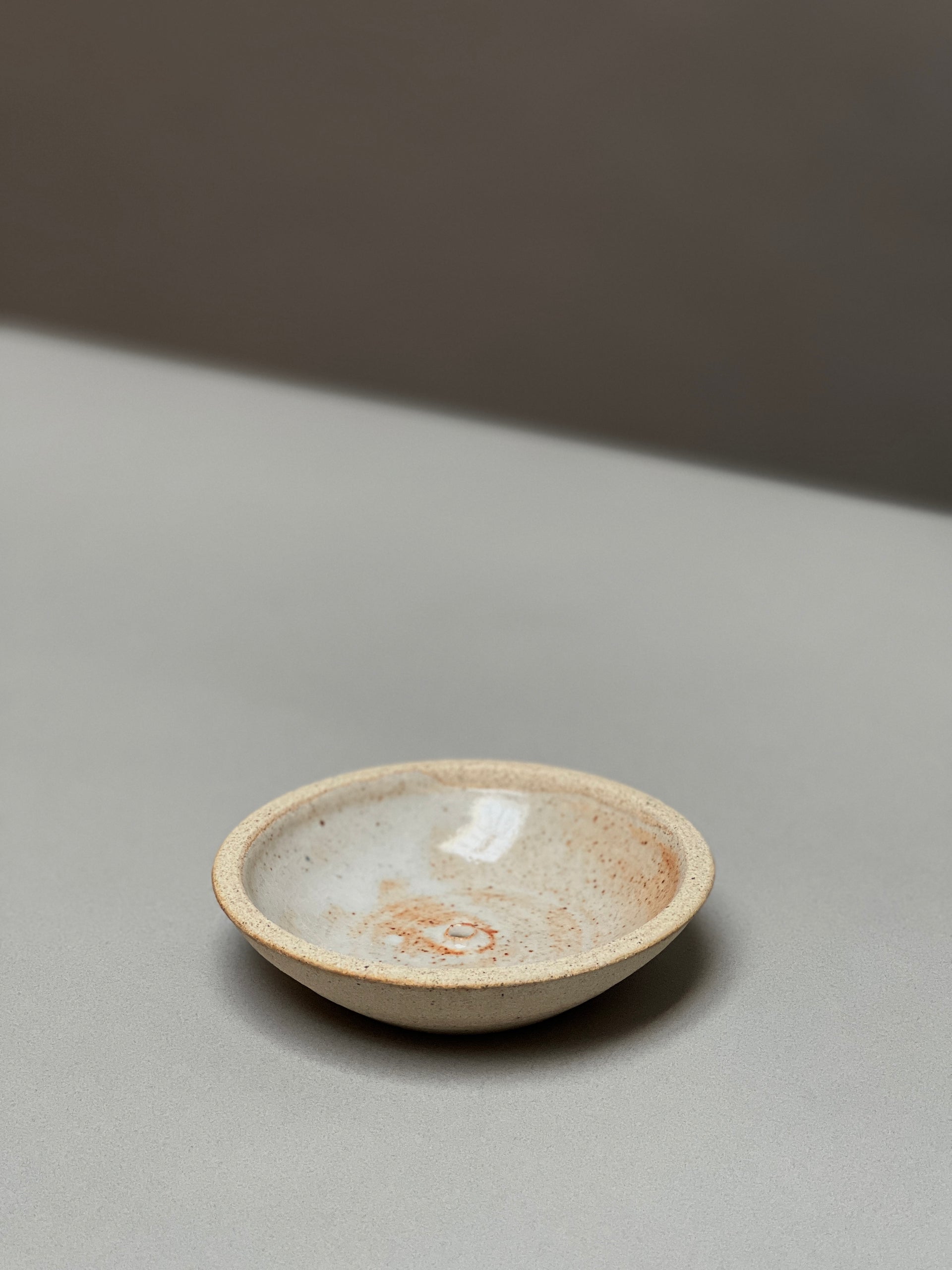 The Stoneware Holder | Incausa by Incausa is a small, shallow hand-thrown incense burner with a light beige and speckled glaze, displayed on a smooth, light gray surface.