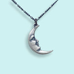 The Silver Man in the Moon Necklace by Ornamental Things features a silver crescent moon pendant with a human face design, suspended from a twisted chain and displayed against a pale blue background—making it a timeless addition to any collection of moon jewelry.