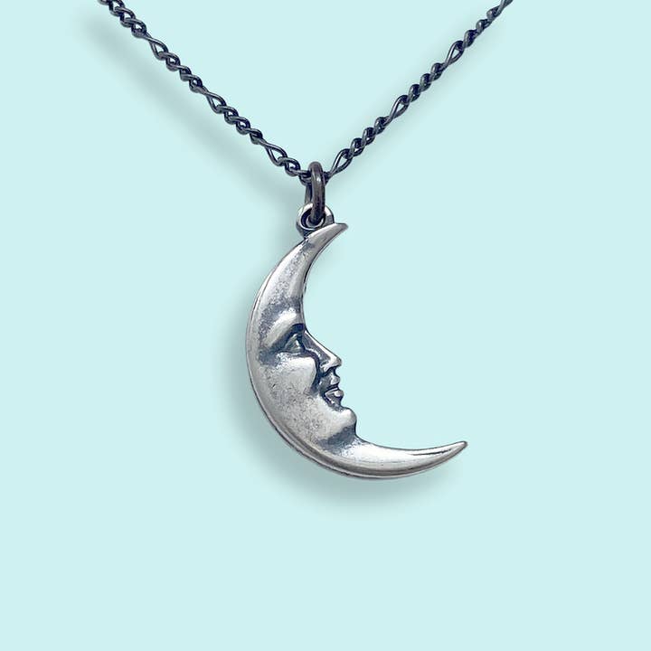 The Silver Man in the Moon Necklace by Ornamental Things features a silver crescent moon pendant with a human face design, suspended from a twisted chain and displayed against a pale blue background—making it a timeless addition to any collection of moon jewelry.