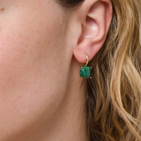 A close-up shows a person’s ear adorned with the JaxKelly Gemstone Mini Drop earrings in malachite—18K gold plated earrings showcasing a square, green gemstone pendant—while light brown hair flows softly in the background.