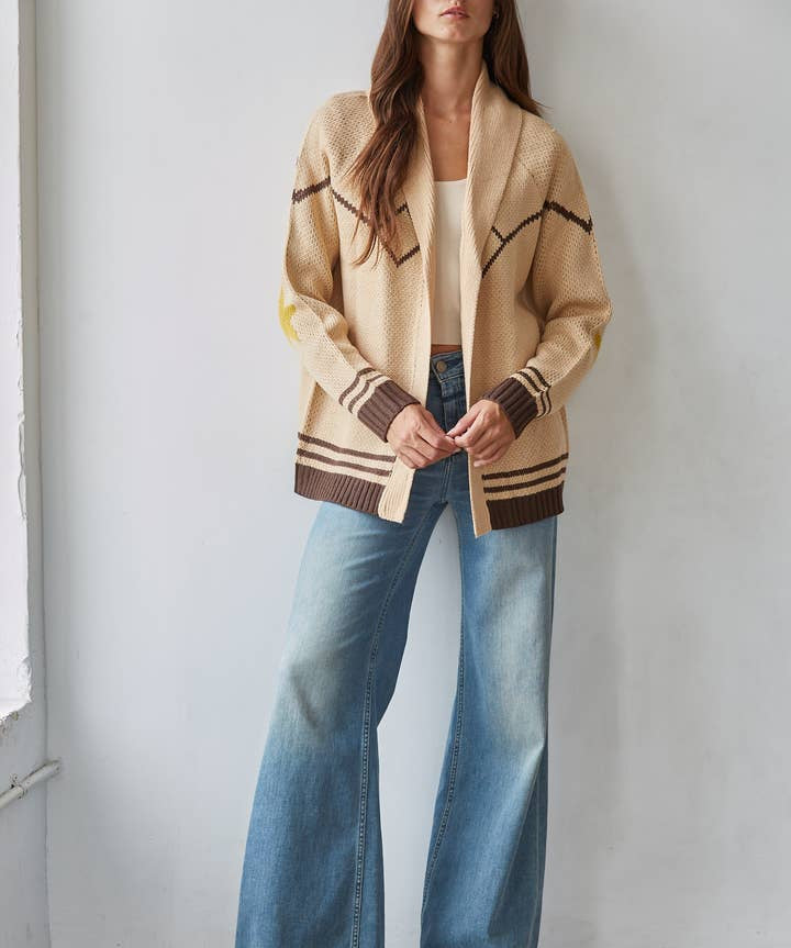 A woman stands against a wall wearing the By Together Knit Sweater Cardigan Top with star and horse jacquard detail, paired with a cream top and high-waisted wide-leg jeans.