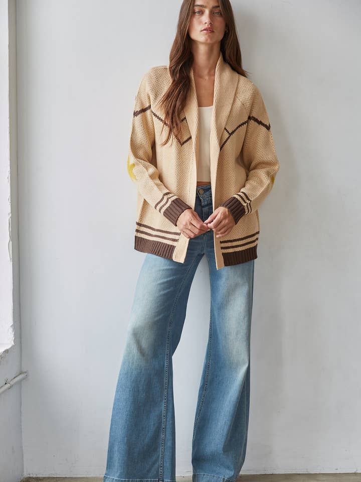 A woman stands against a wall wearing the By Together Knit Sweater Cardigan Top with star and horse jacquard detail, paired with a cream top and high-waisted wide-leg jeans.