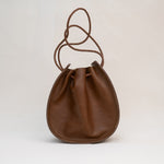 The Joi Bag | Chestnut Distressed from Stash is a Japanese-inspired brown leather drawstring bag featuring a rounded base and a braided, adjustable shoulder strap. Photographed against a plain light background, this handbag blends timeless elegance with practical design.