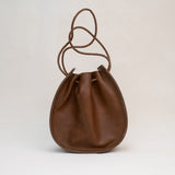 The Joi Bag | Chestnut Distressed from Stash is a Japanese-inspired brown leather drawstring bag featuring a rounded base and a braided, adjustable shoulder strap. Photographed against a plain light background, this handbag blends timeless elegance with practical design.