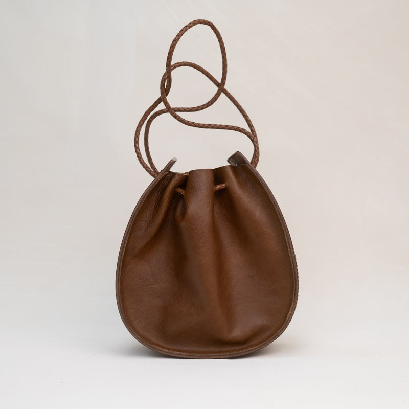 The Joi Bag | Chestnut Distressed from Stash is a Japanese-inspired brown leather drawstring bag featuring a rounded base and a braided, adjustable shoulder strap. Photographed against a plain light background, this handbag blends timeless elegance with practical design.