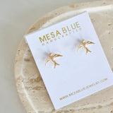 A pair of Mesa Blue Swallow Stud Earrings, elegantly crafted with 18K gold plating, displayed on a white Mesa Blue Handcrafted card atop a textured beige surface.