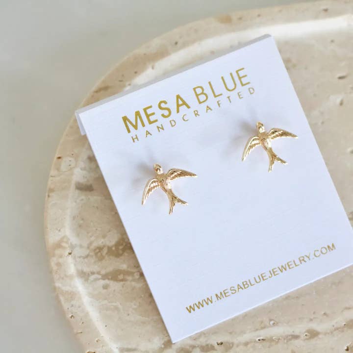 A pair of Mesa Blue Swallow Stud Earrings, elegantly crafted with 18K gold plating, displayed on a white Mesa Blue Handcrafted card atop a textured beige surface.