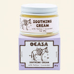 A glass jar of Lavender Tallow Face Cream | OCASA sitting atop its purple cardboard box, both featuring a drawing of a donkey carrying a basket of flowers and text identifying the soothing cream.
