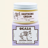 A glass jar of Lavender Tallow Face Cream | OCASA sitting atop its purple cardboard box, both featuring a drawing of a donkey carrying a basket of flowers and text identifying the soothing cream.