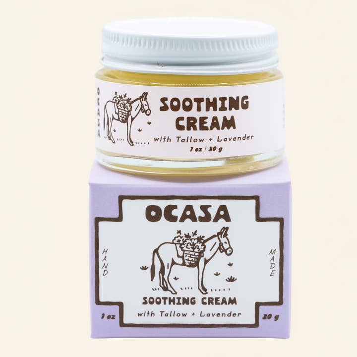 A glass jar of Lavender Tallow Face Cream | OCASA sitting atop its purple cardboard box, both featuring a drawing of a donkey carrying a basket of flowers and text identifying the soothing cream.