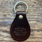 A brown Ballad of the Bird Dog Ballad Leather Keychain, featuring a brass ring and snap and embossed with the words “BALLAD OF THE BAD DOG,” rests on a wooden surface.