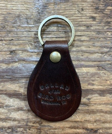 A brown Ballad of the Bird Dog Ballad Leather Keychain, featuring a brass ring and snap and embossed with the words “BALLAD OF THE BAD DOG,” rests on a wooden surface.