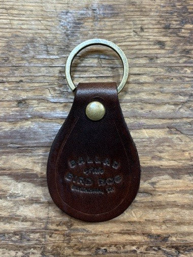 A brown Ballad of the Bird Dog Ballad Leather Keychain, featuring a brass ring and snap and embossed with the words “BALLAD OF THE BAD DOG,” rests on a wooden surface.