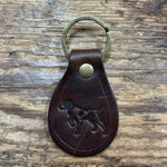 The Ballad of the Bird Dog Ballad Leather Keychain by Ballad of the Bird Dog features a brass ring and an embossed dog image inspired by the Ballad of the Bird Dog, displayed on a wooden surface.