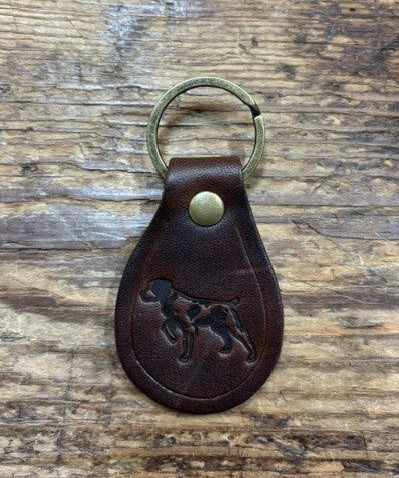 The Ballad of the Bird Dog Ballad Leather Keychain by Ballad of the Bird Dog features a brass ring and an embossed dog image inspired by the Ballad of the Bird Dog, displayed on a wooden surface.