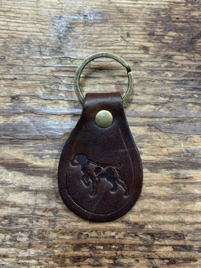 The Ballad of the Bird Dog Ballad Leather Keychain by Ballad of the Bird Dog features a brass ring and an embossed dog image inspired by the Ballad of the Bird Dog, displayed on a wooden surface.