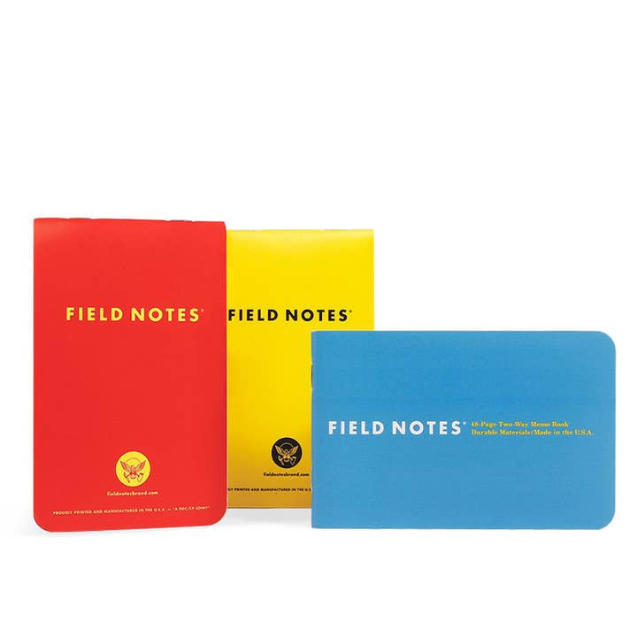 Three Field Notes 1943 Two-way Memo Books, in red, yellow, and blue covers, stand upright side by side against a white background. Inspired by Armed Services Editions, these compact memo books are from Field Notes.