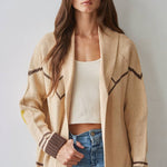 A woman stands against a light wall, wearing the By Together Knit Sweater Cardigan Top with star and horse jacquard detail in beige and brown, layered over a cream crop top and blue jeans.