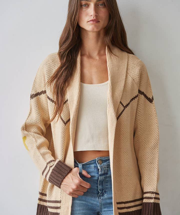 A woman stands against a light wall, wearing the By Together Knit Sweater Cardigan Top with star and horse jacquard detail in beige and brown, layered over a cream crop top and blue jeans.