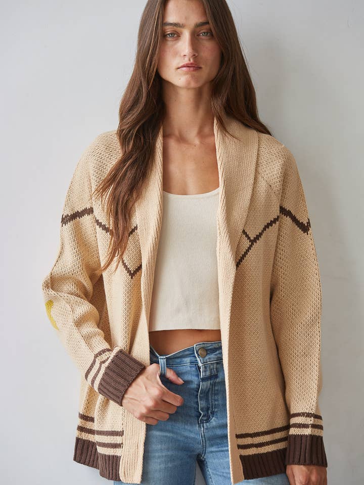 A woman stands against a light wall, wearing the By Together Knit Sweater Cardigan Top with star and horse jacquard detail in beige and brown, layered over a cream crop top and blue jeans.