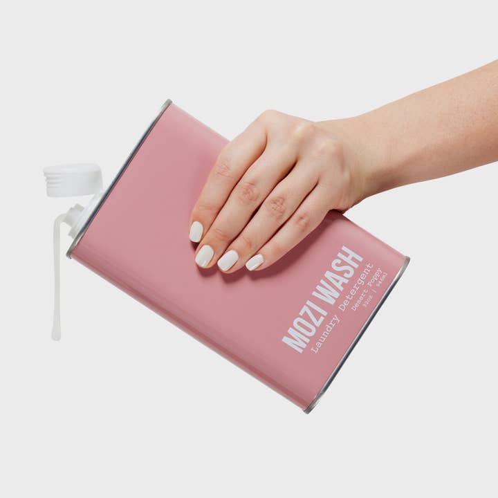 A hand with white nail polish pours liquid from a pink rectangular container labeled “Mozi Desert Poppy Laundry Detergent” with a white cap, releasing a subtle floral fragrance that infuses your laundry with the fresh, uplifting essence of Desert Poppy.