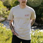 A person with tattoos stands outdoors by a stream, wearing the Hand Tied Flies T-Shirt from Sendero Provisions Co.—a light-colored organic cotton tee featuring “Sendero’s Hand Tied Flies” in red screen print text on the left chest.