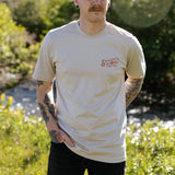 A person with tattoos stands outdoors by a stream, wearing the Hand Tied Flies T-Shirt from Sendero Provisions Co.—a light-colored organic cotton tee featuring “Sendero’s Hand Tied Flies” in red screen print text on the left chest.