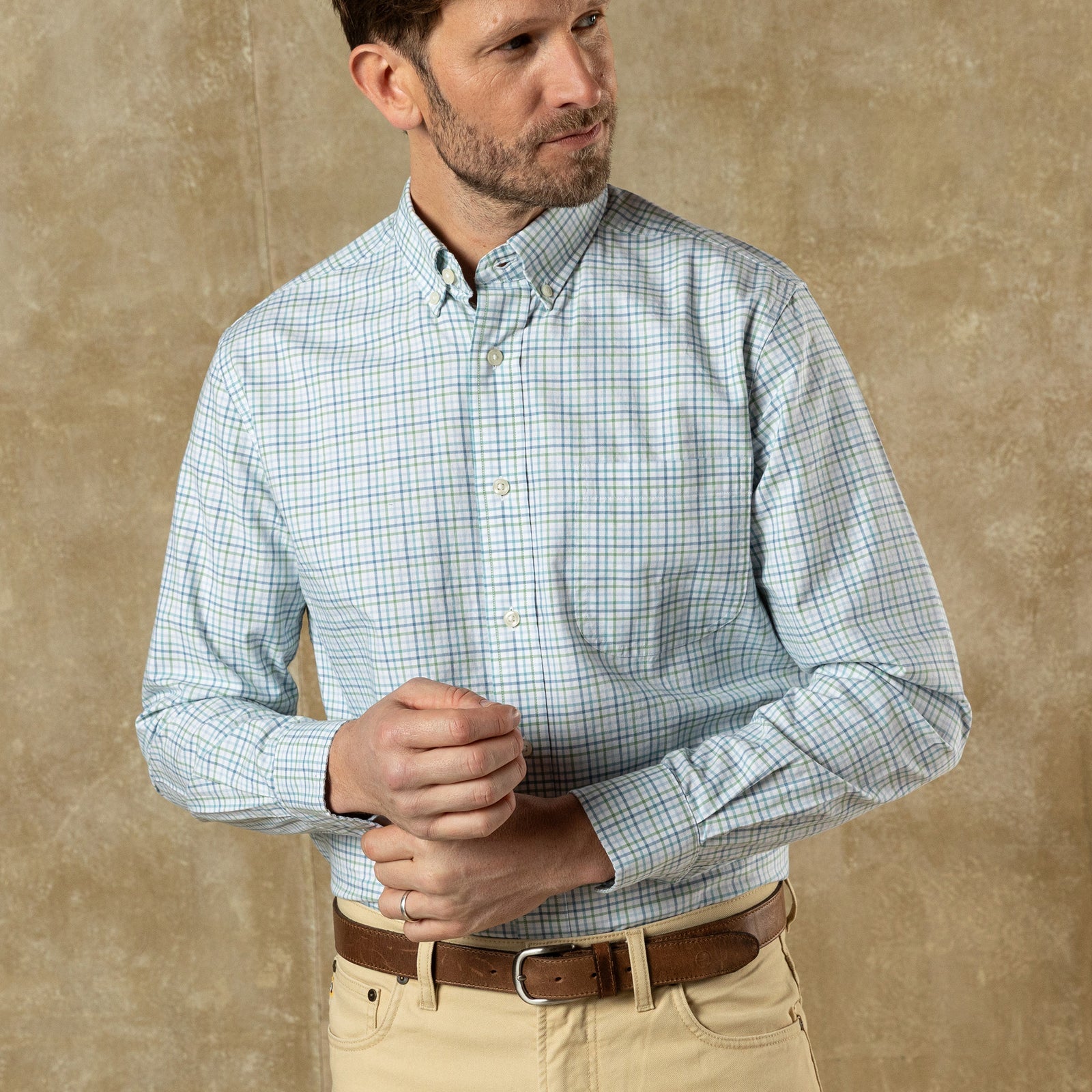 A man adjusts his left cuff while wearing the Duck Head Cotton Oxford Sport Shirt and beige pants, standing against a plain tan background.