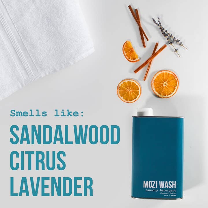 A bottle of Central Coast Laundry Detergent by Mozi is pictured alongside sliced oranges, cinnamon sticks, and sprigs of lavender on a white background. The text reads: “Smells like: Sandalwood Citrus Lavender.” A soft white towel rests in the corner, adding to the refreshing scene.