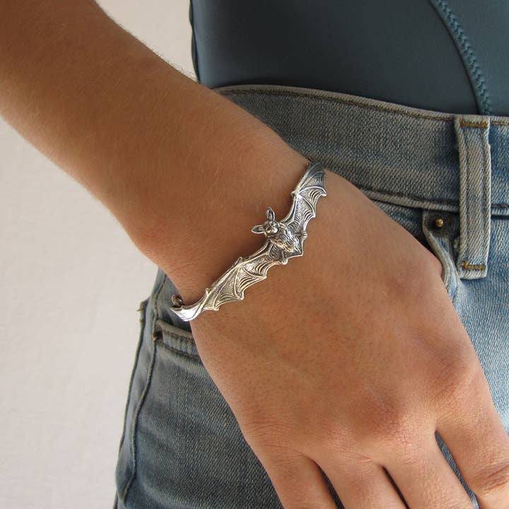 A person wearing high-waisted jeans and the Ornamental Things Silver Bat Bracelet—a vintage-inspired piece shaped like a bat with outstretched wings—on their wrist.
