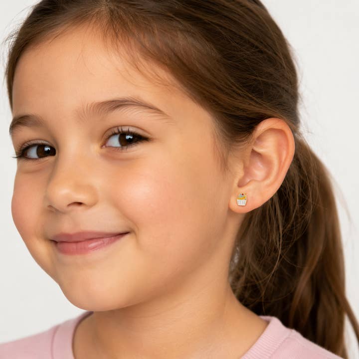 A young girl with brown hair, wearing a light pink shirt and JaxKelly’s Cupcake Enamel Flat Back Kids Earrings, smiles softly against a plain light background—these hypoallergenic earrings are perfect for sensitive ears.