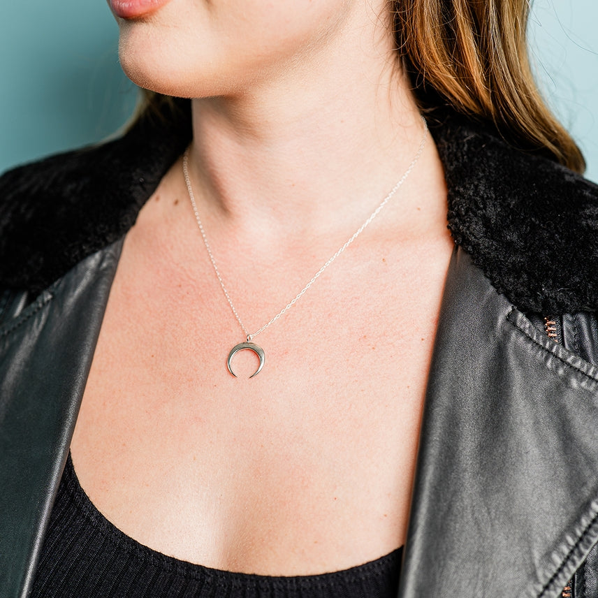 A woman wearing the Sterling Silver Crescent Moon Necklace by Ornamental Things, a black ribbed top, and a black faux fur-collared leather jacket.
