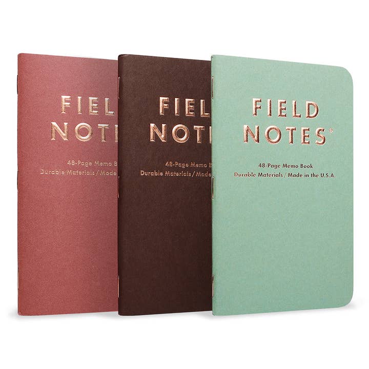 The Field Notes Lucky 3 Pack displays three 48-page memo books with red, brown, and green covers inspired by the classic copper coin, all standing upright in a row.
