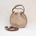 The Joi Bag | Sand Roughout by Stash is a round, beige suede handbag featuring brown braided handles and a detachable, adjustable strap placed in front—this Japanese-inspired accessory blends elegance and versatility for your everyday style.