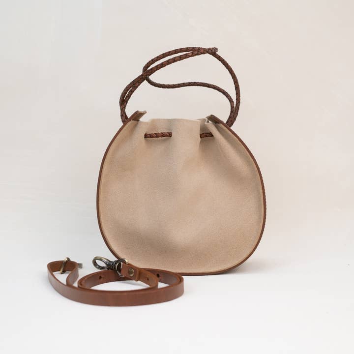 The Joi Bag | Sand Roughout by Stash is a round, beige suede handbag featuring brown braided handles and a detachable, adjustable strap placed in front—this Japanese-inspired accessory blends elegance and versatility for your everyday style.
