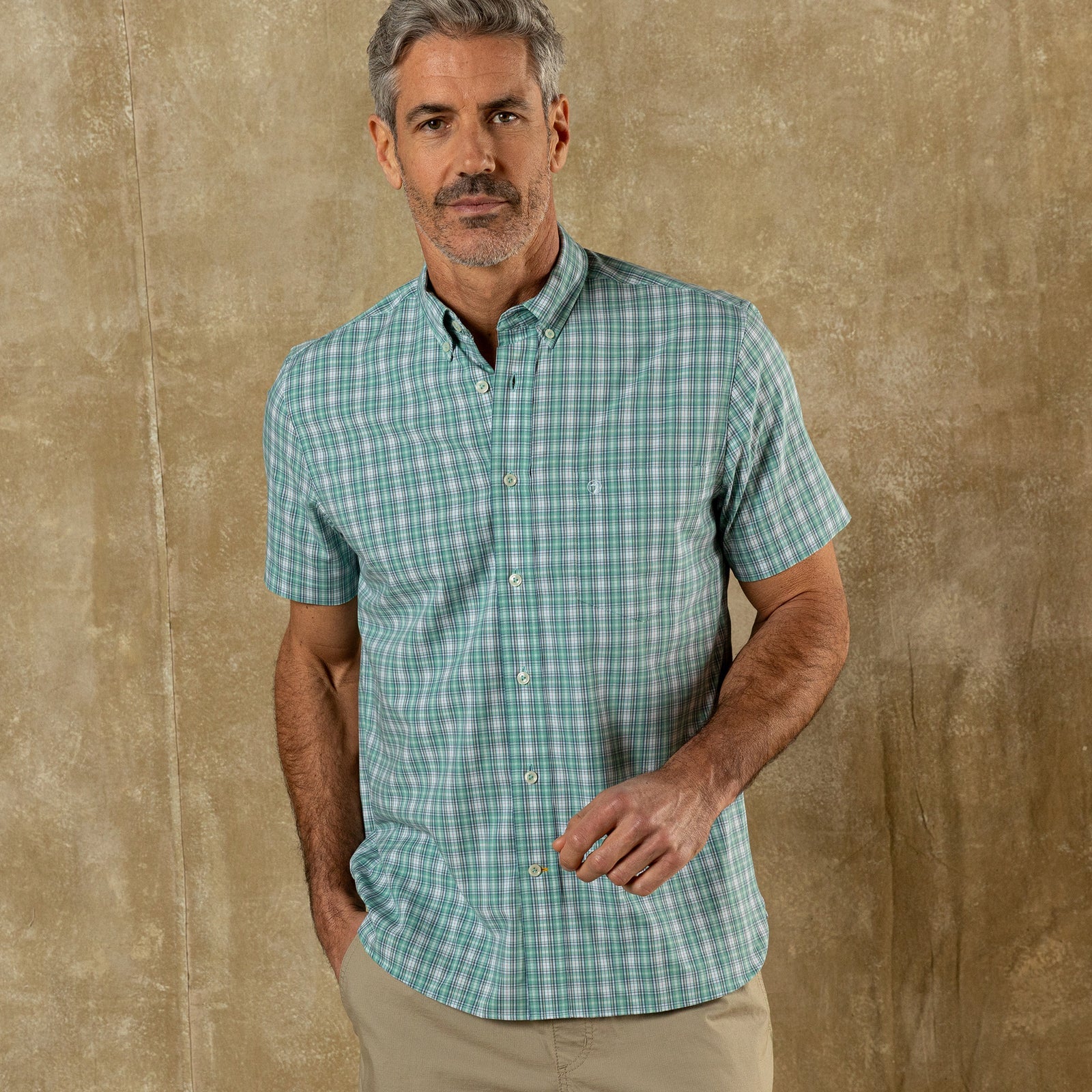 A man with gray hair and a beard wears the Duck Head Performance Poplin Sport Shirt in green plaid and khaki pants, standing against a textured beige background.