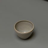 The Stoneware Smudge Bowl by Incausa, a small, round, light gray hand-thrown piece with an in-house glaze, is displayed on a smooth gray surface with minimal shadows.