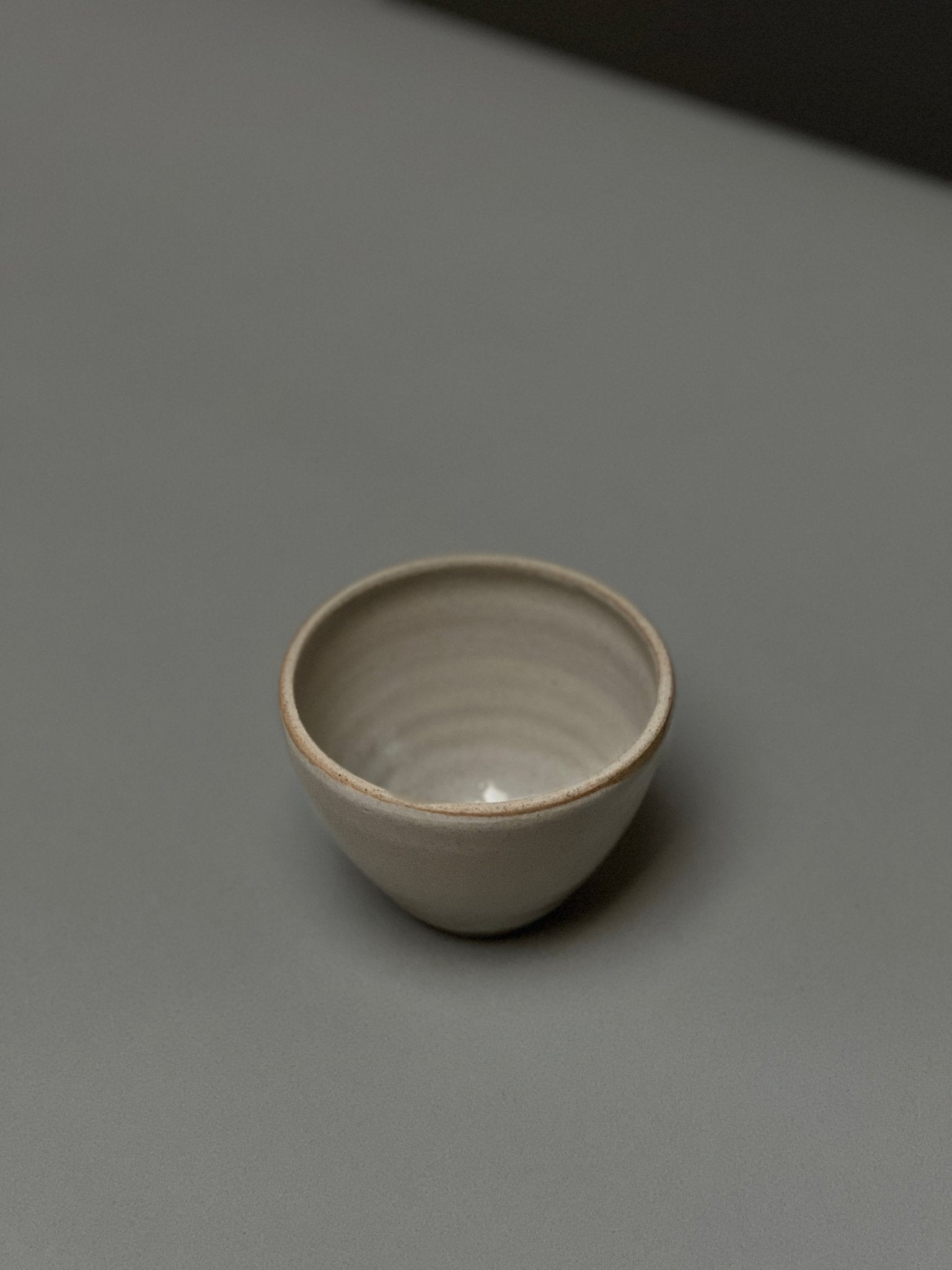 The Stoneware Smudge Bowl by Incausa, a small, round, light gray hand-thrown piece with an in-house glaze, is displayed on a smooth gray surface with minimal shadows.
