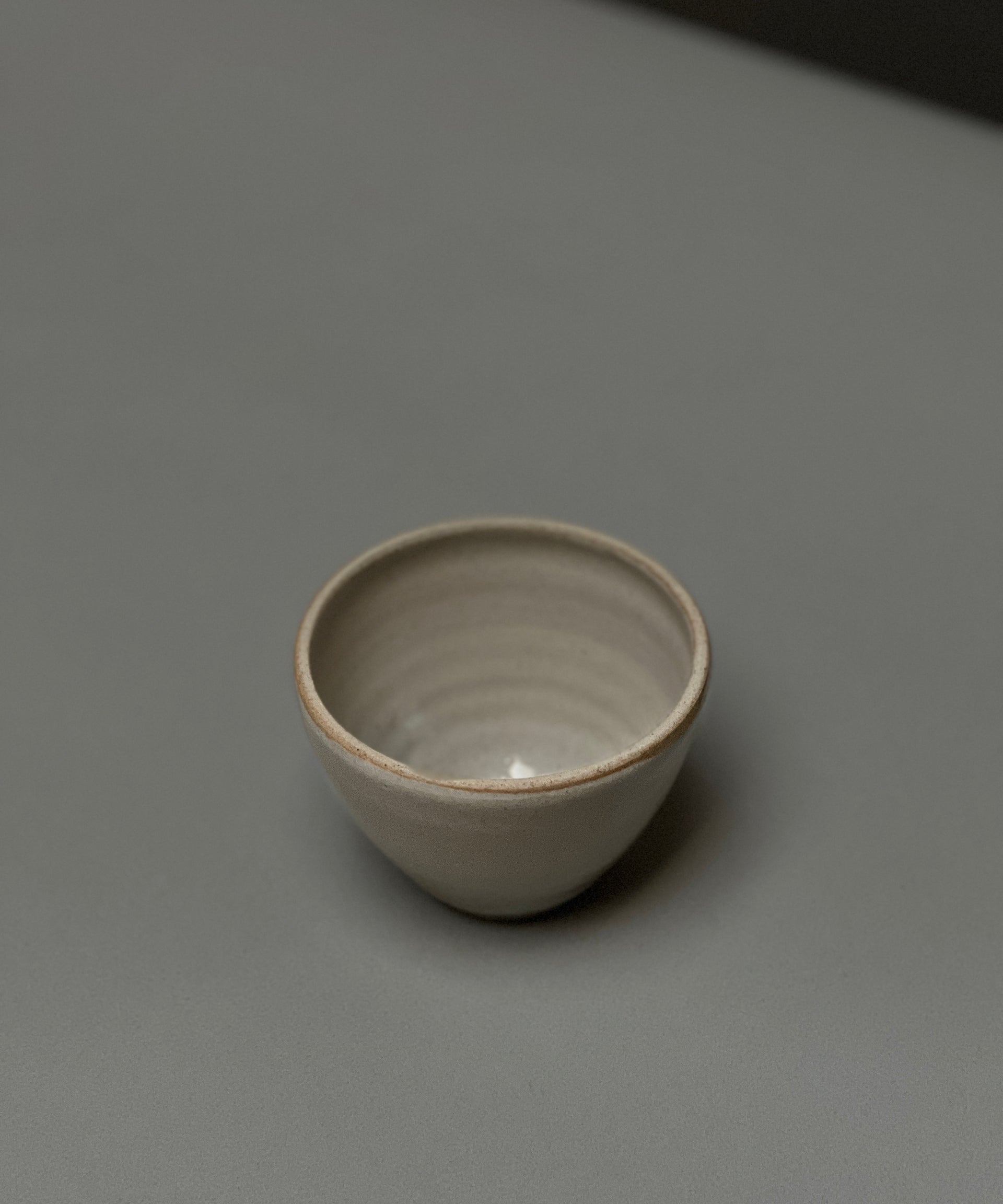 The Stoneware Smudge Bowl by Incausa, a small, round, light gray hand-thrown piece with an in-house glaze, is displayed on a smooth gray surface with minimal shadows.