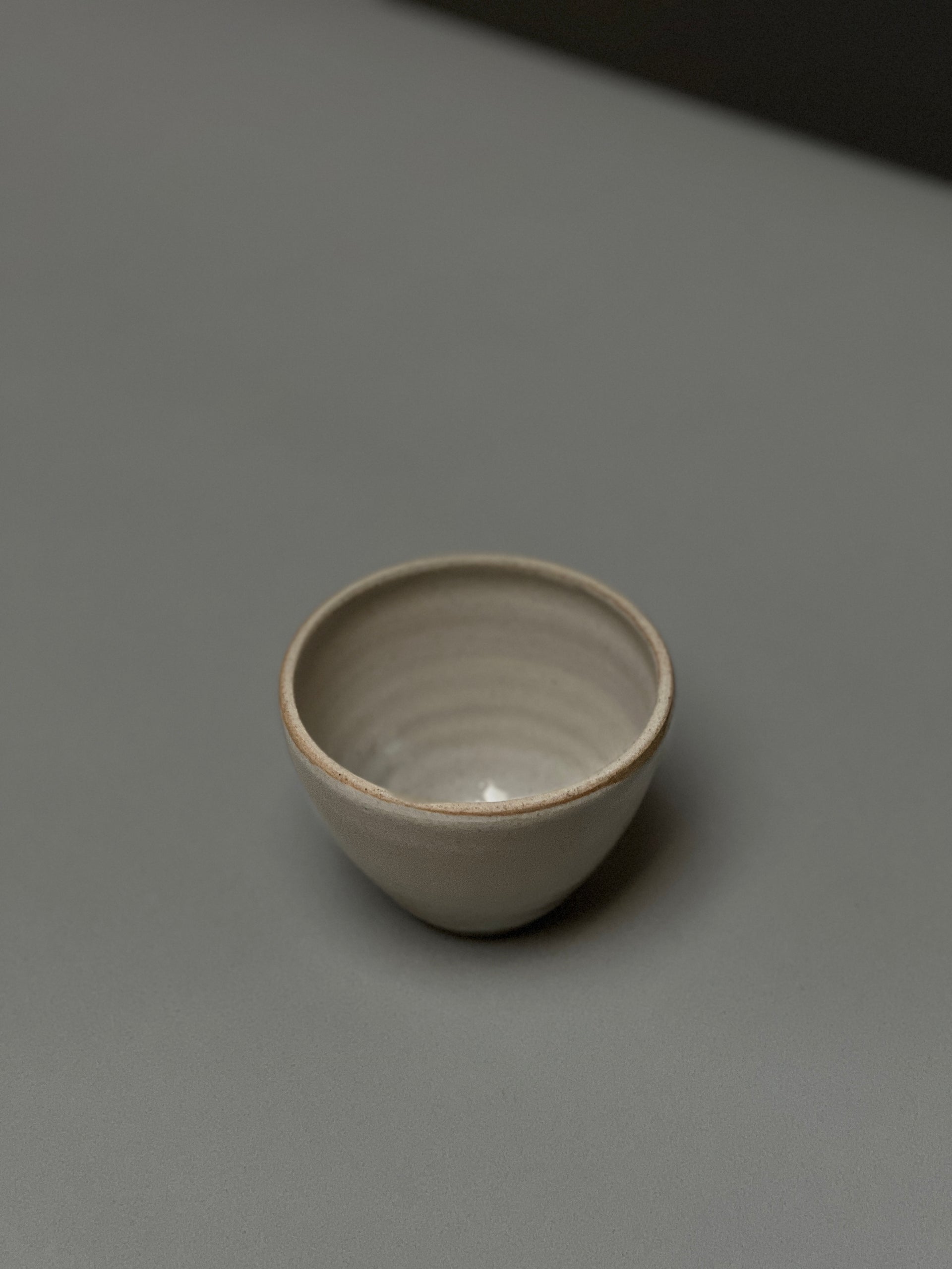 The Stoneware Smudge Bowl by Incausa, a small, round, light gray hand-thrown piece with an in-house glaze, is displayed on a smooth gray surface with minimal shadows.
