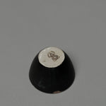 The Incausa Stoneware Smudge Bowl is shown upside down on a gray surface, displaying its beige base with a dark geometric design and in-house glaze.