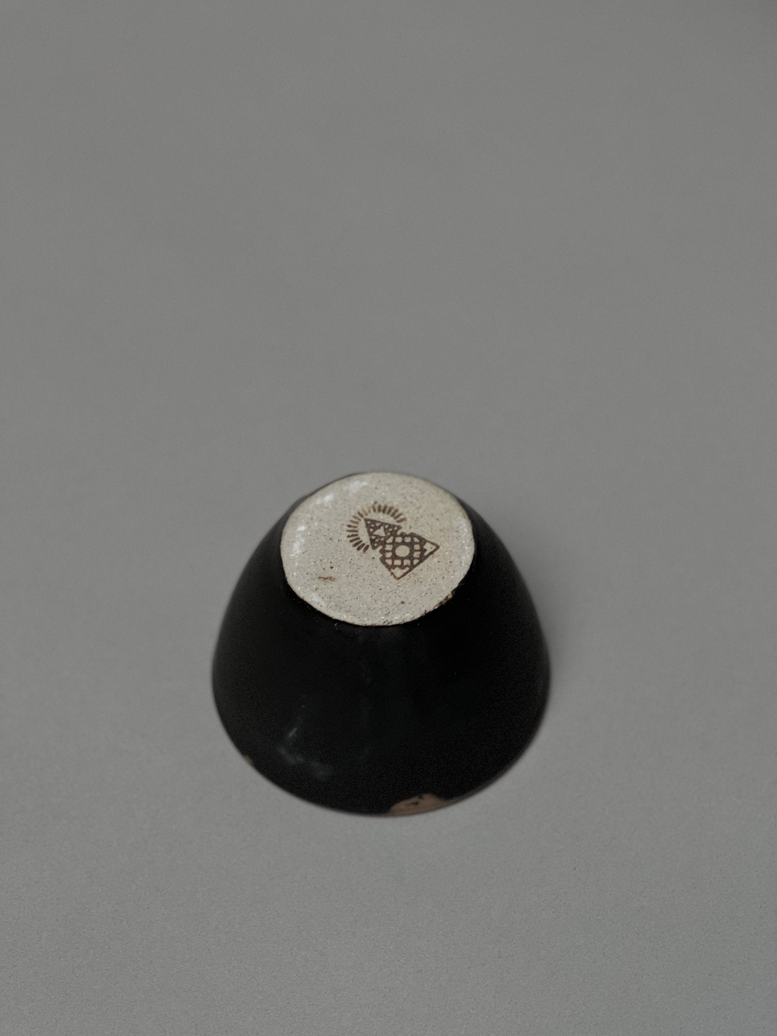 The Incausa Stoneware Smudge Bowl is shown upside down on a gray surface, displaying its beige base with a dark geometric design and in-house glaze.