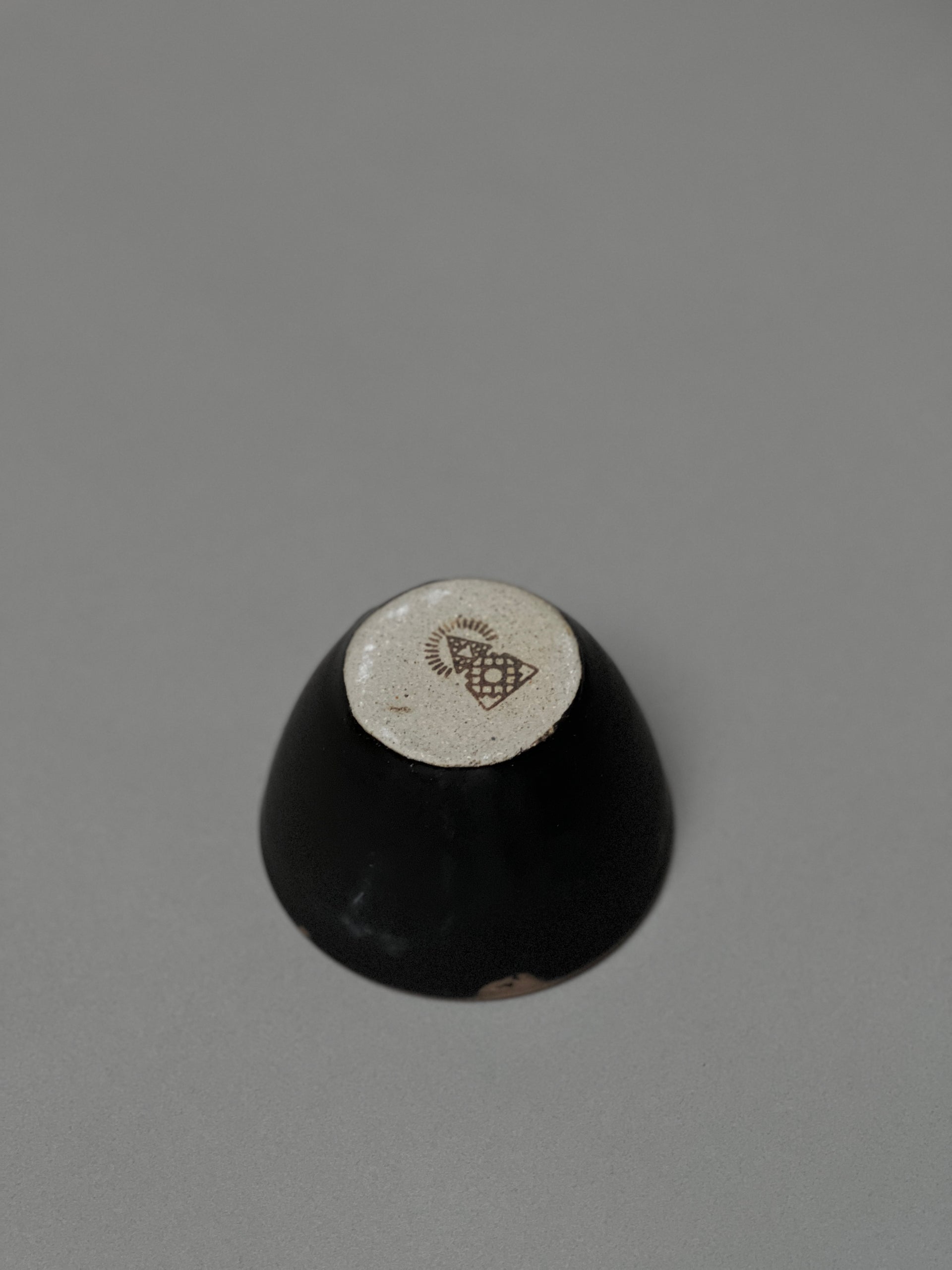 The Incausa Stoneware Smudge Bowl is shown upside down on a gray surface, displaying its beige base with a dark geometric design and in-house glaze.