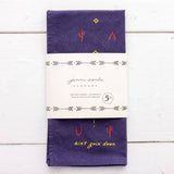 The Heritage Cotton Bandana - “Ain’T Goin’ Down” by Jenni Earle is a folded purple western bandana featuring small red and yellow designs, with the phrase “ain’t goin’ down” in yellow. It comes wrapped in a white paper label with Jenni Earle branding and product details—an ideal encouragement gift or a stylish cotton accessory for any occasion.