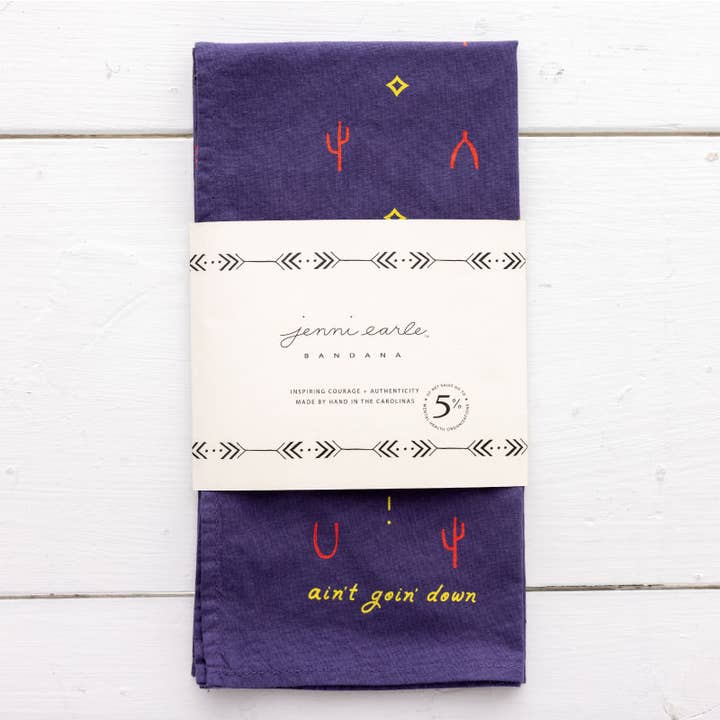 The Heritage Cotton Bandana - “Ain’T Goin’ Down” by Jenni Earle is a folded purple western bandana featuring small red and yellow designs, with the phrase “ain’t goin’ down” in yellow. It comes wrapped in a white paper label with Jenni Earle branding and product details—an ideal encouragement gift or a stylish cotton accessory for any occasion.