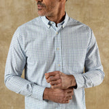 A man wearing the Duck Head Cotton Twill Sport Shirt with a button-down collar in light blue and white check stands against a beige textured background, adjusting his cuff.