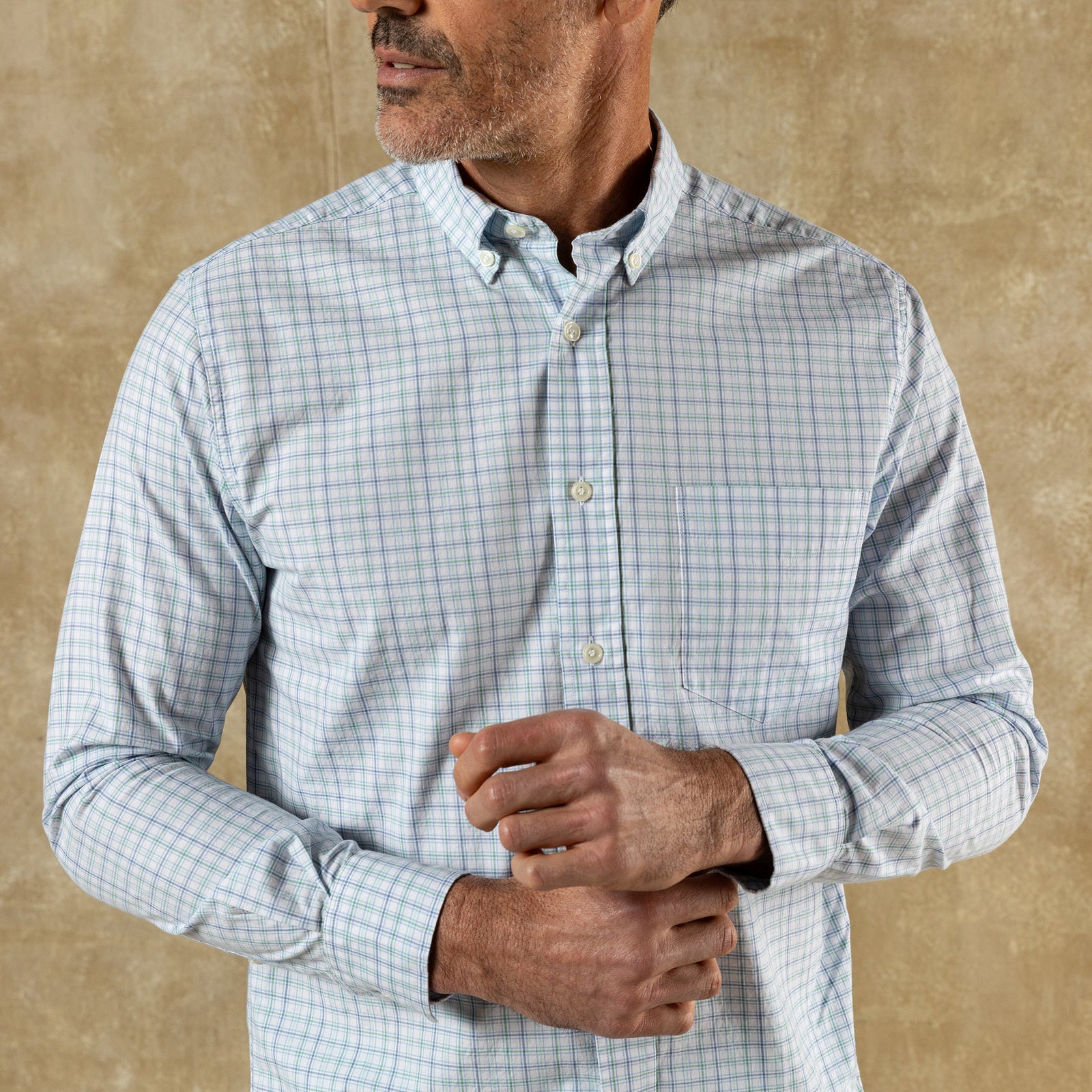 A man wearing the Duck Head Cotton Twill Sport Shirt with a button-down collar in light blue and white check stands against a beige textured background, adjusting his cuff.