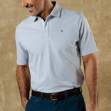 A man stands against a tan textured background wearing Duck Head’s Hanover Pima Cotton Pique Polo in light blue, tucked into blue pants with a brown belt, exemplifying classic style.